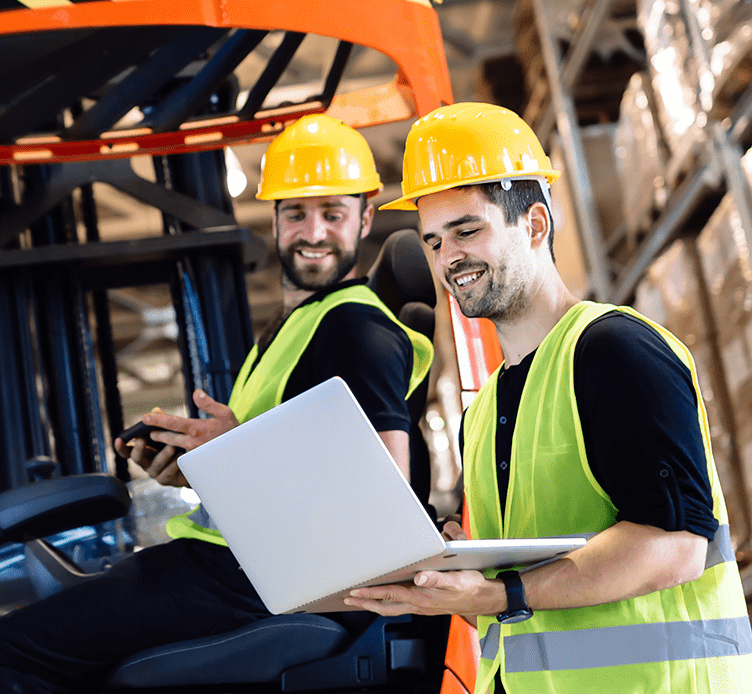 Two men with hard hats and safety vests looking at a laptop in a warehouse.